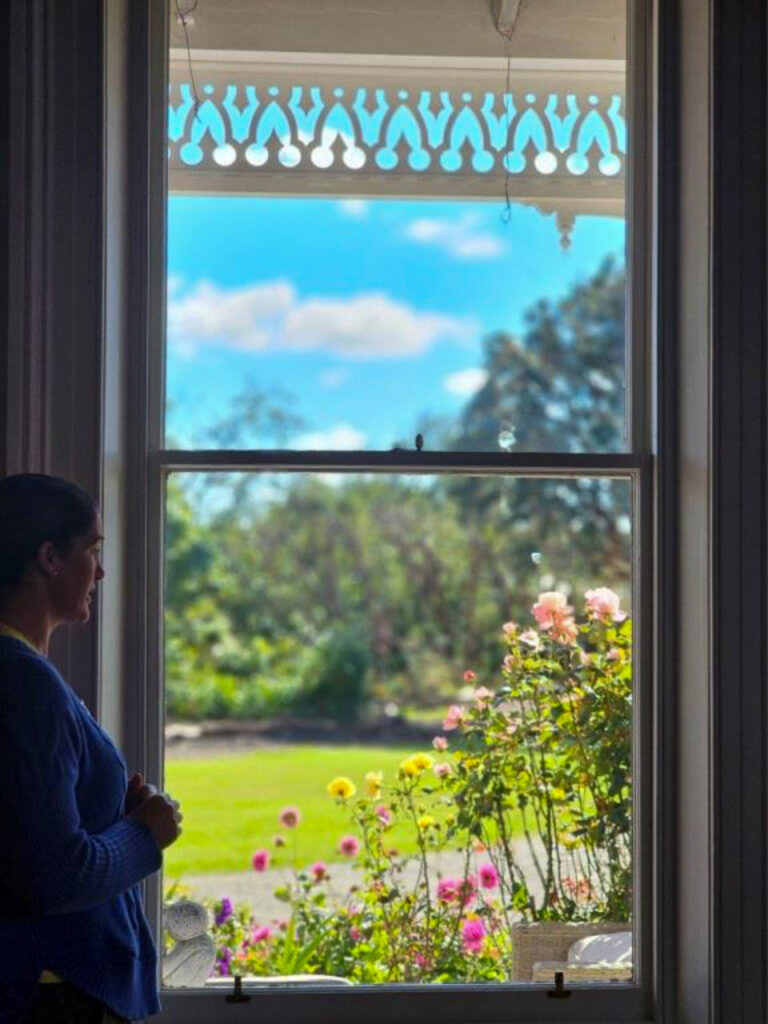 Laura Thompson at Lennel House window looking at the beautiful garden with trees and flowers.